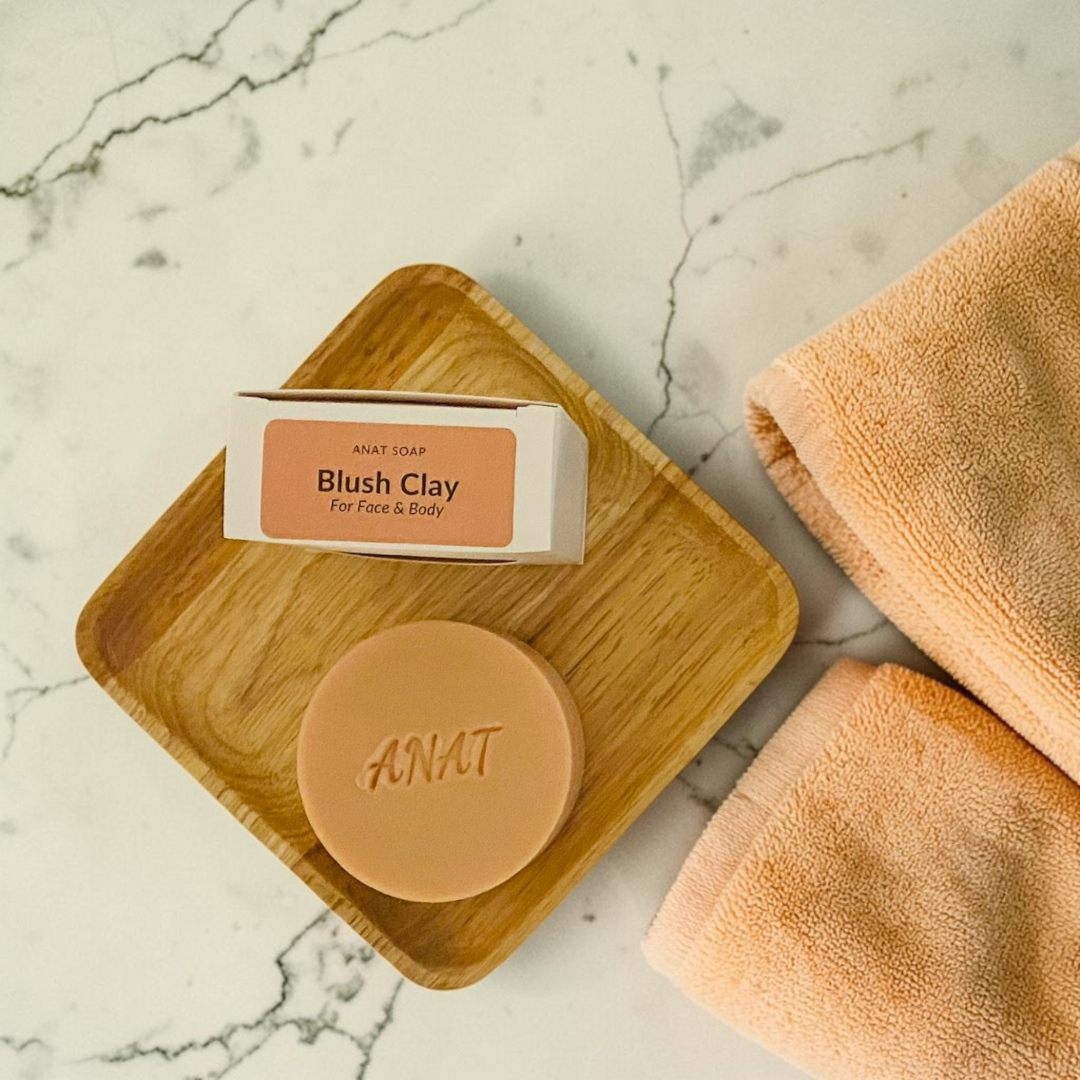 Anat Soap Blush Clay round handmade plant-based soap bar with mango and kokum butters for face and body, shown on a bamboo soap dish next to the product box.