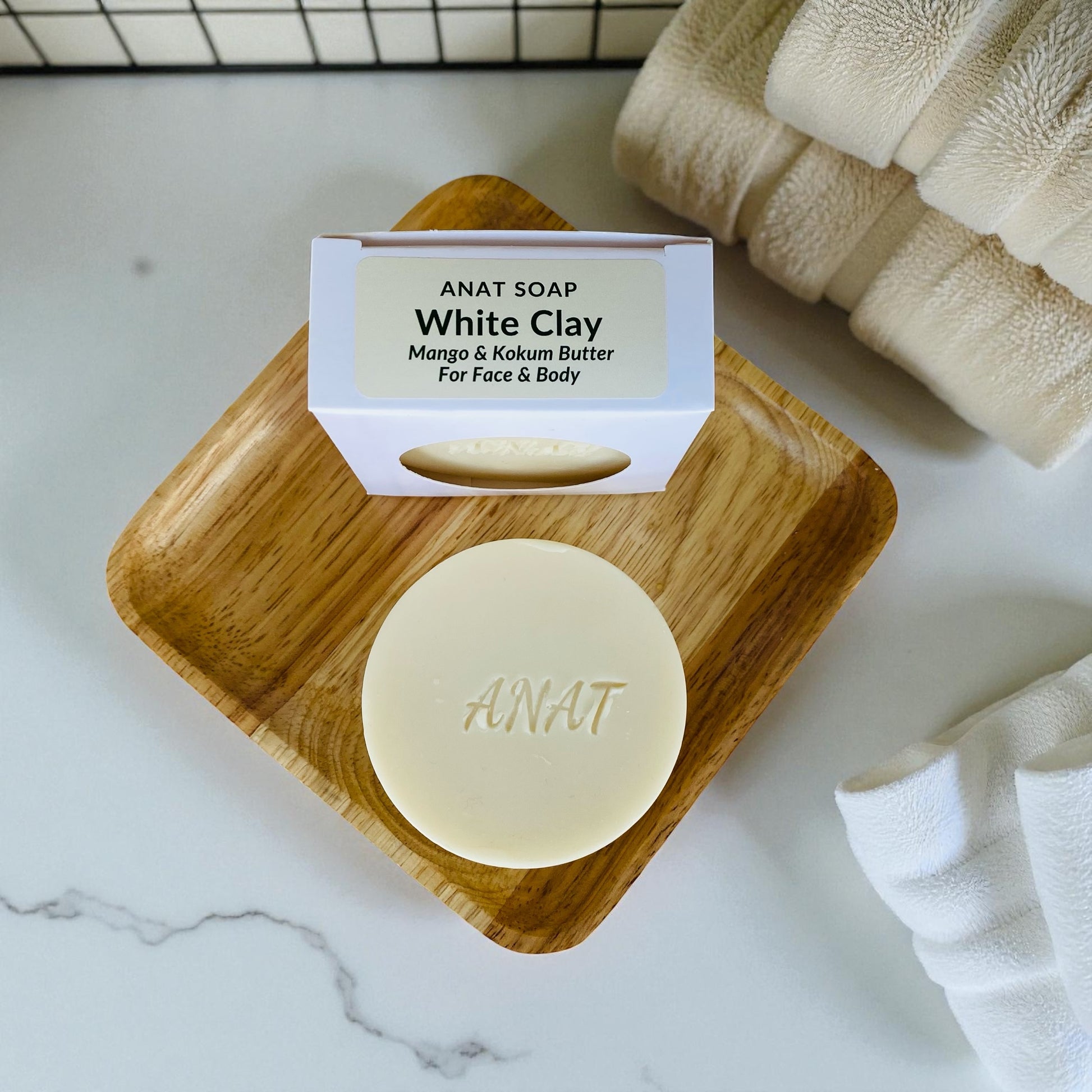 Anat Soap White Clay round handmade plant-based soap bar with mango and kokum butters for face and body, shown on a bamboo soap dish next to the product box.